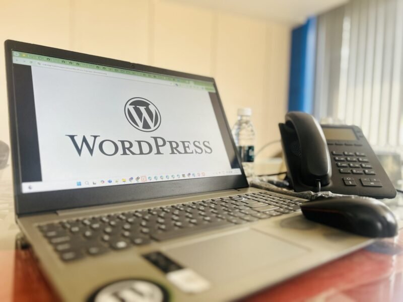 Laptop displaying WordPress on an office desk with a smartphone and desk phone.